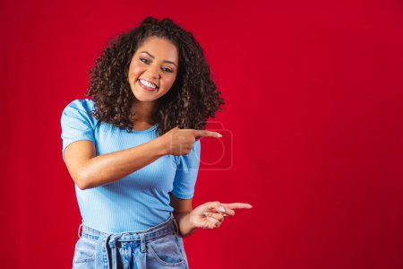 Photo for Cheerful Afro woman points away on copy space, discusses amazing promo, gives way or direction, wears yellow warm sweater, has pleasant smile, feels optimistic, isolated over red background. - Royalty Free Image