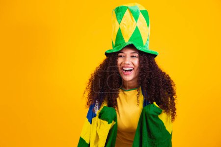 Photo for Afro girl cheering for favorite brazilian team, holding national flag in yellow background. - Royalty Free Image