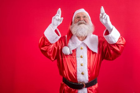Photo for Portrait of dreamy elderly man in santa claus costume  crossing fingers making a wish, christmas magic, winter holidays. Indoor studio shot isolated on red background - Royalty Free Image