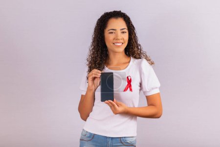 Téléchargez les photos : Le concept de la Journée mondiale du sida. Une jeune femme tient un smartphone avec un écran vierge et un ruban rouge sur un t-shirt avec un concept de prévention et de planification du VIH. - en image libre de droit