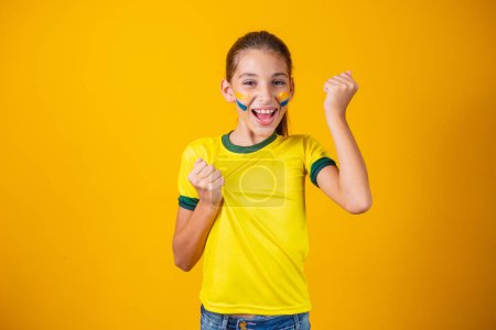 Téléchargez les photos : Supporteur de football, équipe du Brésil. Belle petite fille acclamant pour son équipe sur fond jaune - en image libre de droit