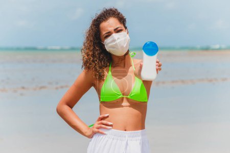 Photo for Afro woman in bikini and mask holding sunscreen bottle in hand on the beach. - Royalty Free Image