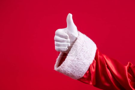 Foto de Primer plano de la mano con guante de Santa Claus con el pulgar hacia arriba haciendo señal de ok sobre fondo rojo. Feliz Año Nuevo, Feliz Navidad, tradicional celebración estacional. - Imagen libre de derechos