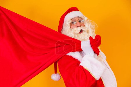 Foto de Santa Claus sobre fondo amarillo sosteniendo bolsa con regalos con espacio libre para el texto a sus espaldas. campañas navideñas - Imagen libre de derechos