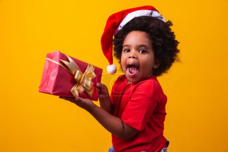 Foto de Sonriente niño afro en Santa Claus sombrero rojo con regalo de Navidad en la mano. Concepto de Navidad. - Imagen libre de derechos