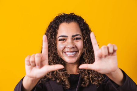 Foto de Chica afroamericana joven con ropa ejecutiva sonriendo haciendo marco fotográfico con manos y dedos con una cara feliz. concepto de creatividad y fotografía. - Imagen libre de derechos