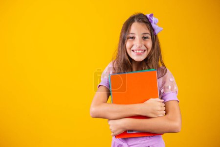 Téléchargez les photos : Belle fille souriante tenant des livres allant à l'école. portrait rapproché, fond jaune isolé, enfance. cahiers de câlins pour enfants. mode de vie, intérêt, passe-temps, temps libre, temps libre - en image libre de droit