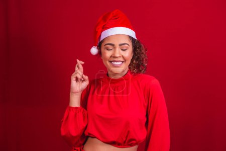 Photo for Young woman in christmas outfit and santa claus hat with fingers crossed wishing luck - Royalty Free Image