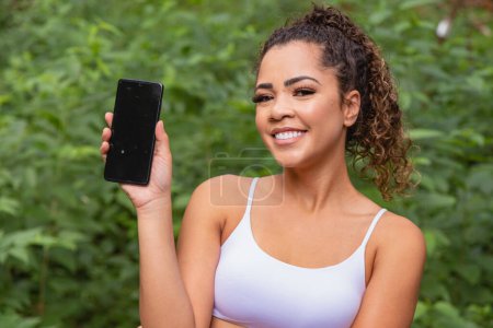 Foto de Aplicación deportiva. Retrato de joven en forma emocionada sosteniendo y mostrando el teléfono inteligente, señalando e indicando una pantalla en blanco para el texto. Exterior - Imagen libre de derechos