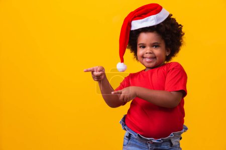 Foto de Pequeña Afro niña en camiseta roja y sombrero de Santa vestida para Navidad con espacio para el texto apuntando. - Imagen libre de derechos
