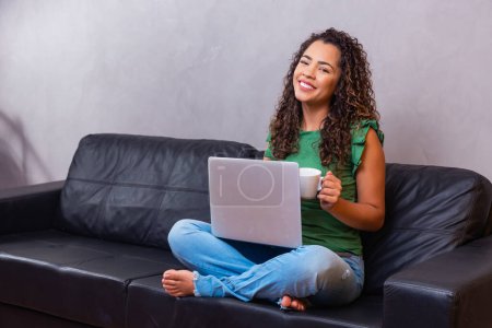 Téléchargez les photos : Sourire afro jeune femme à l'aide d'un ordinateur portable, assis sur le canapé à la maison, belle fille faisant du shopping ou bavarder en ligne dans les réseaux sociaux, s'amuser, regarder un film, pigiste travaillant sur un projet informatique - en image libre de droit