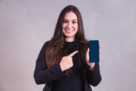Téléchargez les photos : Femme caucasienne pointant vers le téléphone portable avec de l'espace pour le texte sur fond gris. - en image libre de droit
