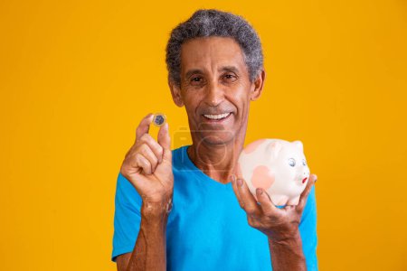 Foto de Hombre mayor sosteniendo una alcancía y una moneda brasileña sobre fondo amarillo. concepto de financiación - Imagen libre de derechos