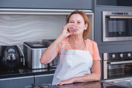 Téléchargez les photos : Femme mûre boire de l'eau dans la cuisine le matin - en image libre de droit