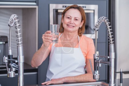 Foto de Mujer madura bebiendo agua en la cocina por la mañana - Imagen libre de derechos