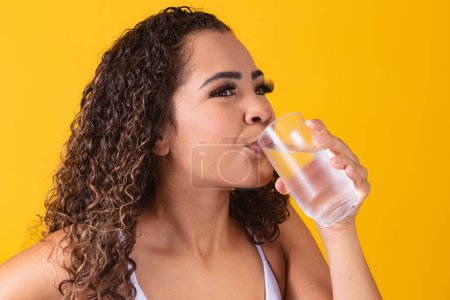 Téléchargez les photos : Jeune afro femme boire de l'eau en fond jaune. Jeune fille avec un verre d'eau. Jeune femme buvant de l'eau du verre. Concept d'hydratation - en image libre de droit