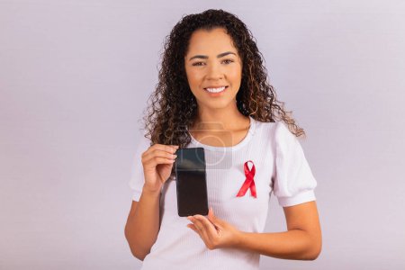 Téléchargez les photos : Le concept de la Journée mondiale du sida. Une jeune femme tient un smartphone avec un écran vierge et un ruban rouge sur un t-shirt avec un concept de prévention et de planification du VIH. - en image libre de droit