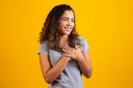 Foto de Mujer africana feliz en camiseta gris pose sobre fondo amarillo sonriendo mirando a la cámara cogida de la mano en el pecho sintiendo gratitud, corazón, sentimientos sinceros gesto y concepto de amor - Imagen libre de derechos