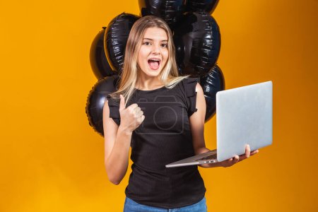 Foto de Sonriente alegre divertida joven morena veinteañera utilizando ordenador portátil PC sobre fondo de color amarillo brillante con globos de aire retrato de estudio. Viernes negro venta - Imagen libre de derechos