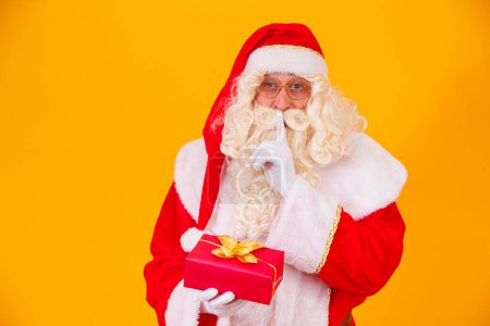 Foto de Santa Claus sobre fondo amarillo sosteniendo un regalo en las manos y haciendo una señal de silencio con la otra mano. Santa Claus sorpresa - Imagen libre de derechos