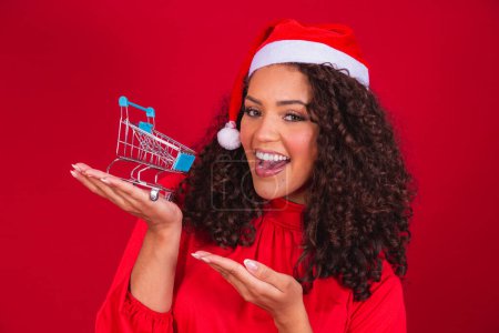 Photo for Afro Woman in Santa hat doing christmas shopping. She is showing a mini cart - Royalty Free Image