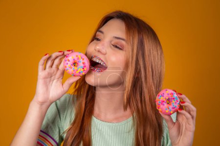 Foto de Chica pelirroja sobre fondo amarillo comiendo deliciosas rosquillas coloridas. - Imagen libre de derechos