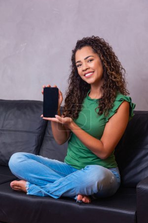 Photo for Afro woman sitting on sofa holding cellphone with white screen with free space for text. - Royalty Free Image