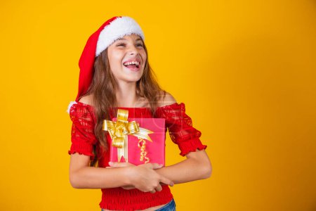 Téléchargez les photos : Mignonne jolie fille gaie en chapeau de Noël sur fond coloré tenant un cadeau - en image libre de droit