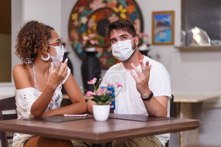 Photo for Couple of friends safety talking sitting on an outdoors restaurant wearing safety mask against the covid-19 coronavirus pandemic. New normal life concept - Royalty Free Image
