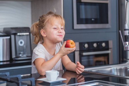 Téléchargez les photos : Petite fille blonde manger une pomme dans la cuisine - en image libre de droit