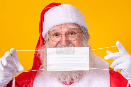 Foto de El verdadero Santa Claus con un fondo amarillo, sosteniendo una máscara protectora, gafas y sombrero. Navidad con distancia social. Covid-19 - Imagen libre de derechos