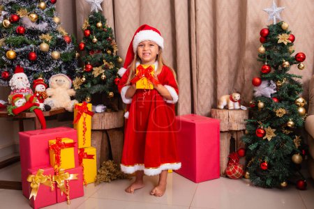 Photo for Cute little caucasian girl dressed for christmas at home holding a gift. - Royalty Free Image
