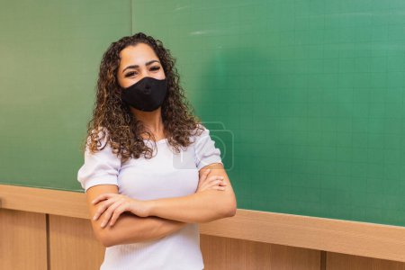 Photo for Young female teacher with arms crossed in classroom with blackboard in background wearing surgical mask in new normal. Concept of back to school after pandemic - Royalty Free Image