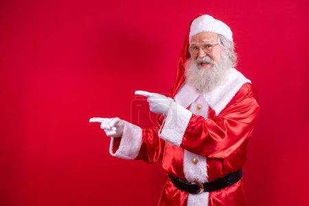 Photo for Funny happy excited old bearded Santa Claus face wearing costume looking at camera showing pointing fingers aside advertising Christmas promotion, New Year xmas discount ad isolated on red background. - Royalty Free Image