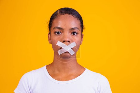 Téléchargez les photos : Femme noire sur fond jaune avec la bouche fermée en silence. Notion de préjudice, d'abus et de racisme - en image libre de droit