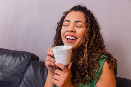 Téléchargez les photos : Jeune femme afro se détendre sur le canapé prendre le thé ou le café d'une tasse. - en image libre de droit
