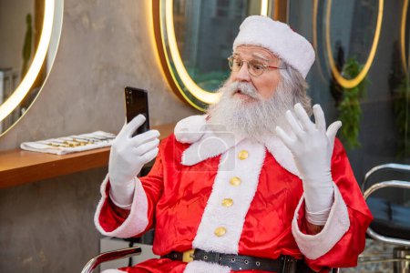 Foto de Santa Claus al teléfono en una videollamada mientras se prepara en el salón de belleza para la víspera de Navidad - Imagen libre de derechos