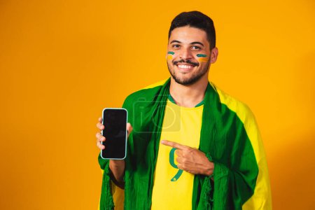Brasilianischer Fan. Porträt, brasilianischer Fan zeigt sein Handy, gekleidet als Fußballfan oder Fußballspiel auf gelbem Hintergrund. Farben Brasiliens. Weltmeisterschaft.