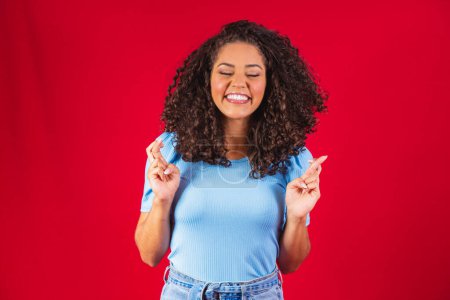 Photo for I have to win. Joyful young brunette female clenches teeth, raises fingers crossed, makes desirable wish, curly hair, mixed race, African, Brazilian girl, waits for good news, stands over red wall - Royalty Free Image
