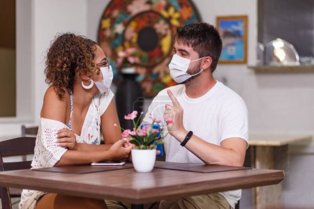Foto de Un par de amigos de seguridad hablando sentados en un restaurante al aire libre con máscara de seguridad contra la pandemia de coronavirus covid-19. Nuevo concepto de vida normal - Imagen libre de derechos