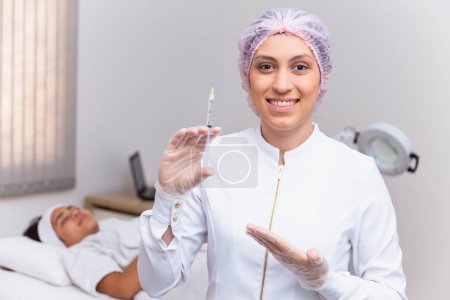 Téléchargez les photos : Femme appliquant de l'acide hyaluronique sur son visage. Esthéticienne tenant une seringue avec de l'acide hyaluronique et patiente en arrière-plan. - en image libre de droit