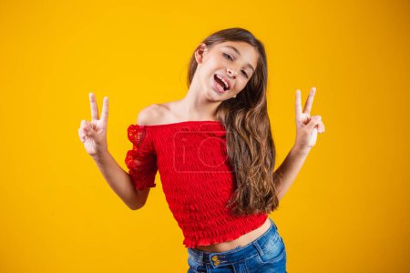 Foto de Foto de una encantadora y delicada niña preadolescente haciendo un signo V sonriendo aislada sobre fondo amarillo. - Imagen libre de derechos