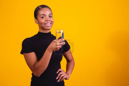 Téléchargez les photos : Jeune afro femme boire de l'eau en fond jaune. Jeune fille avec verre d'eau - en image libre de droit