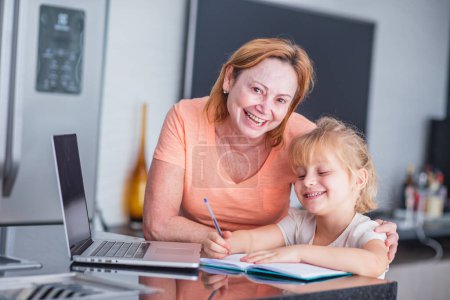 Foto de Sonriente madre madura ayuda a su hija en la preparación de tareas en casa. Concepto de educación en línea. - Imagen libre de derechos
