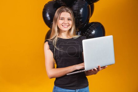 Téléchargez les photos : Souriant joyeux drôle jeune femme brune 20s en utilisant ordinateur portable PC sur fond de couleur jaune vif avec des ballons à air portrait studio. Vente vendredi noir - en image libre de droit
