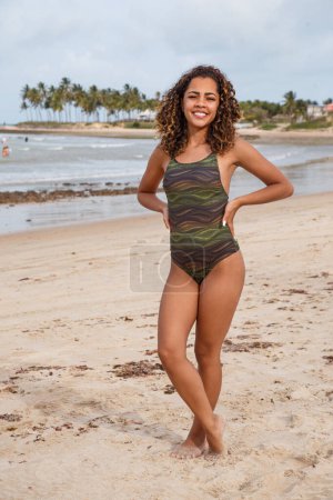 Téléchargez les photos : Belle femme latino-américaine en bikini sur la plage. Jeune femme profitant de ses vacances d'été par une journée ensoleillée, souriant et regardant la caméra - en image libre de droit