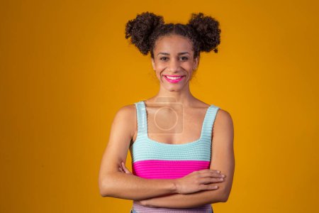 Téléchargez les photos : Afro jeune femme avec les bras croisés sur fond jaune avec un chignon dans les cheveux. - en image libre de droit