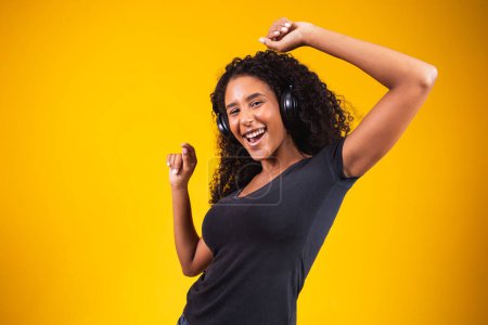 Téléchargez les photos : Jeune femme africaine portant un casque dansant actif isolé sur un mur jaune écoutant de la musique rire gai. - en image libre de droit