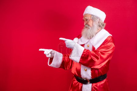 Photo for Santa Claus pointing aside, advertising Christmas promotion, New Year Christmas discount advertisement isolated on red background. - Royalty Free Image