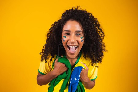 Téléchargez les photos : Afro fille acclamant pour l'équipe brésilienne préférée, tenant drapeau national en arrière-plan jaune
. - en image libre de droit